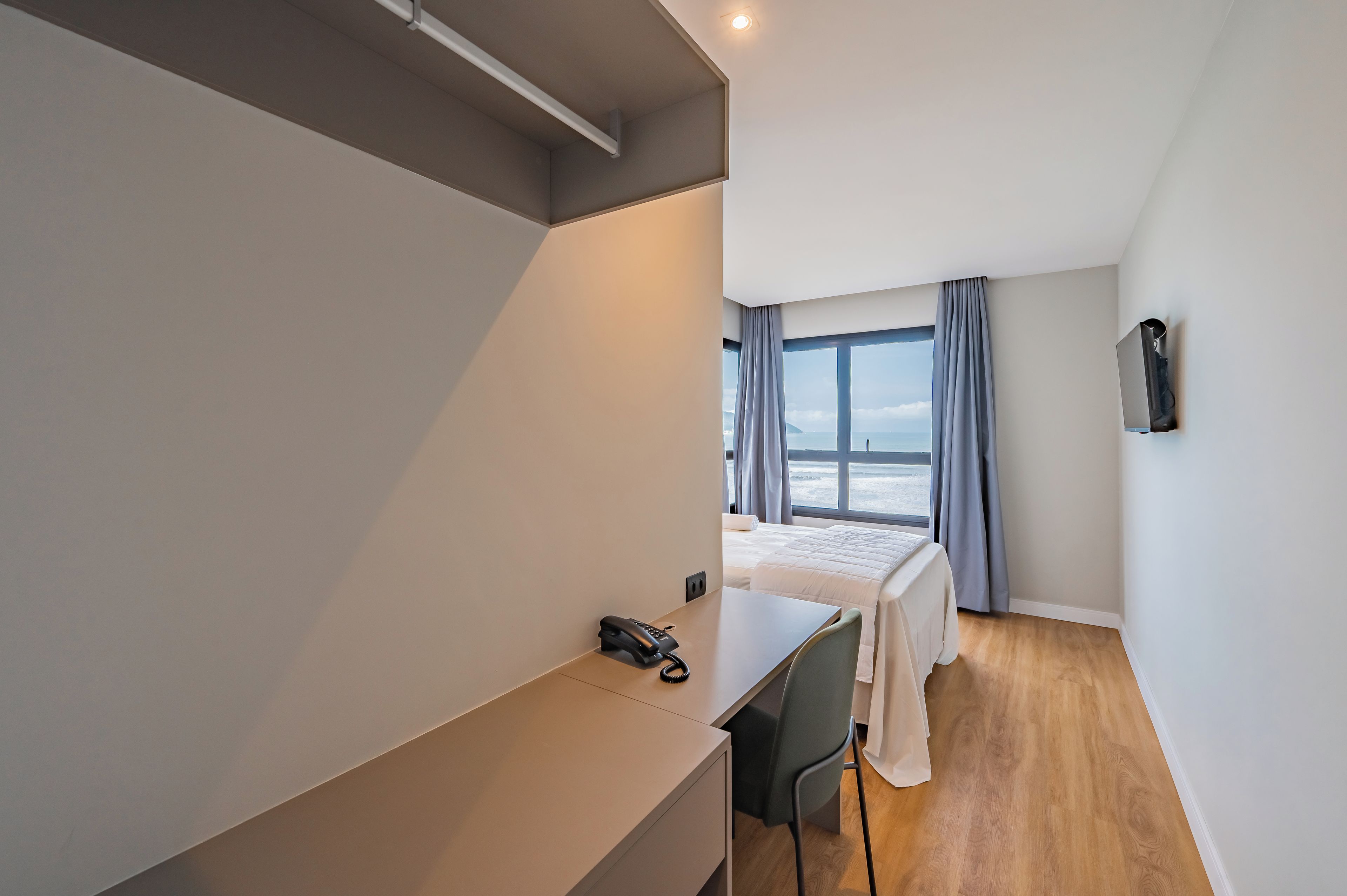 Quarto de hotel com cama, escrivaninha, cadeira, telefone e janela com cortinas.