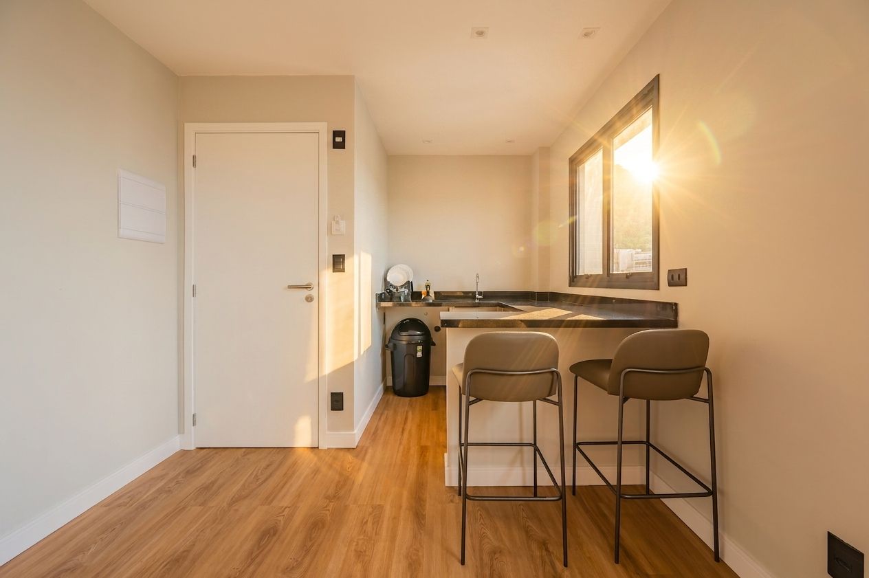Cocina compacta con puerta blanca, una pequeña barra con dos taburetes y un cesto de basura.