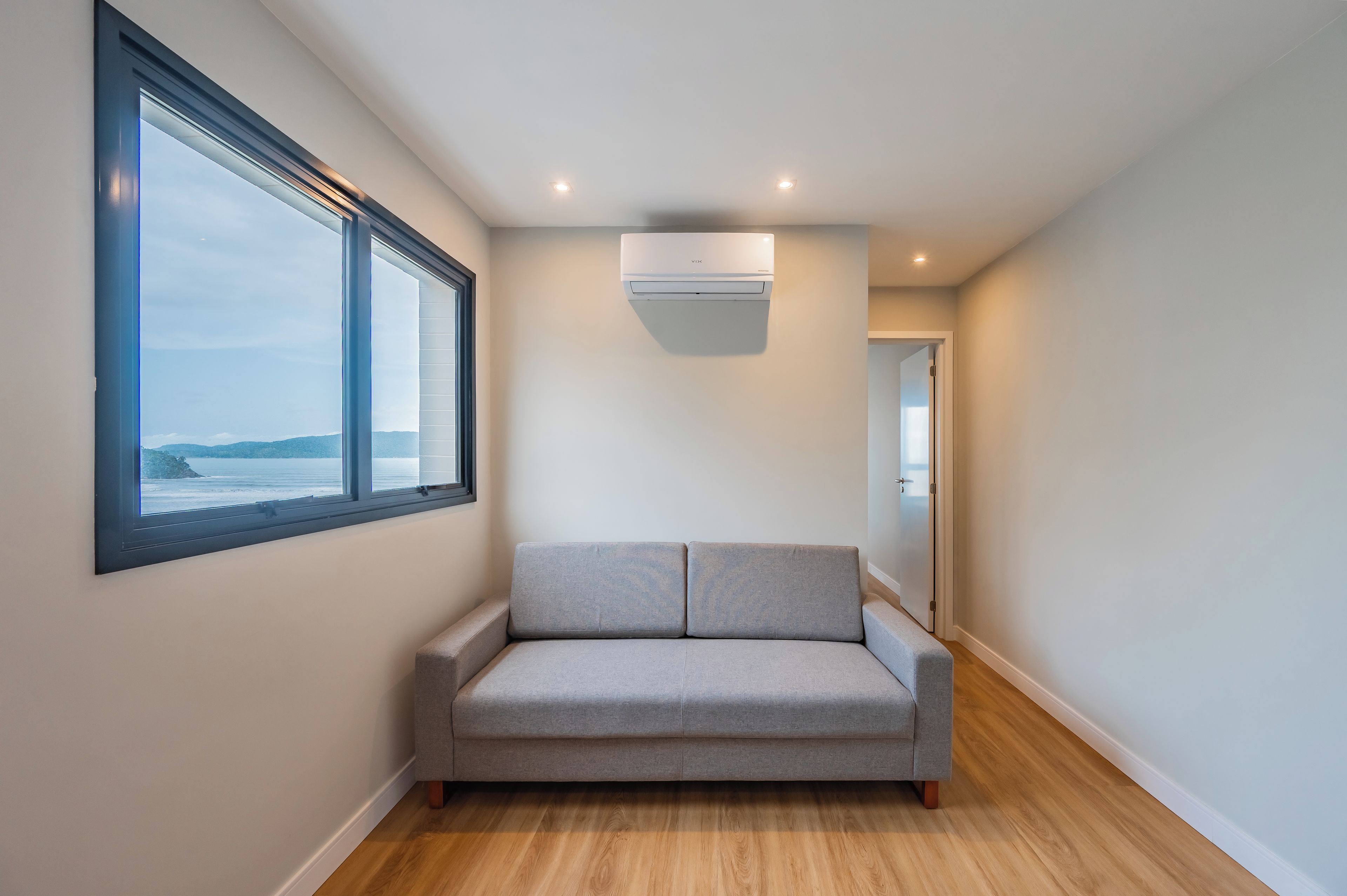 A narrow hallway with a grey sofa, a window with a view of the sea, and an air conditioning unit on the wall.