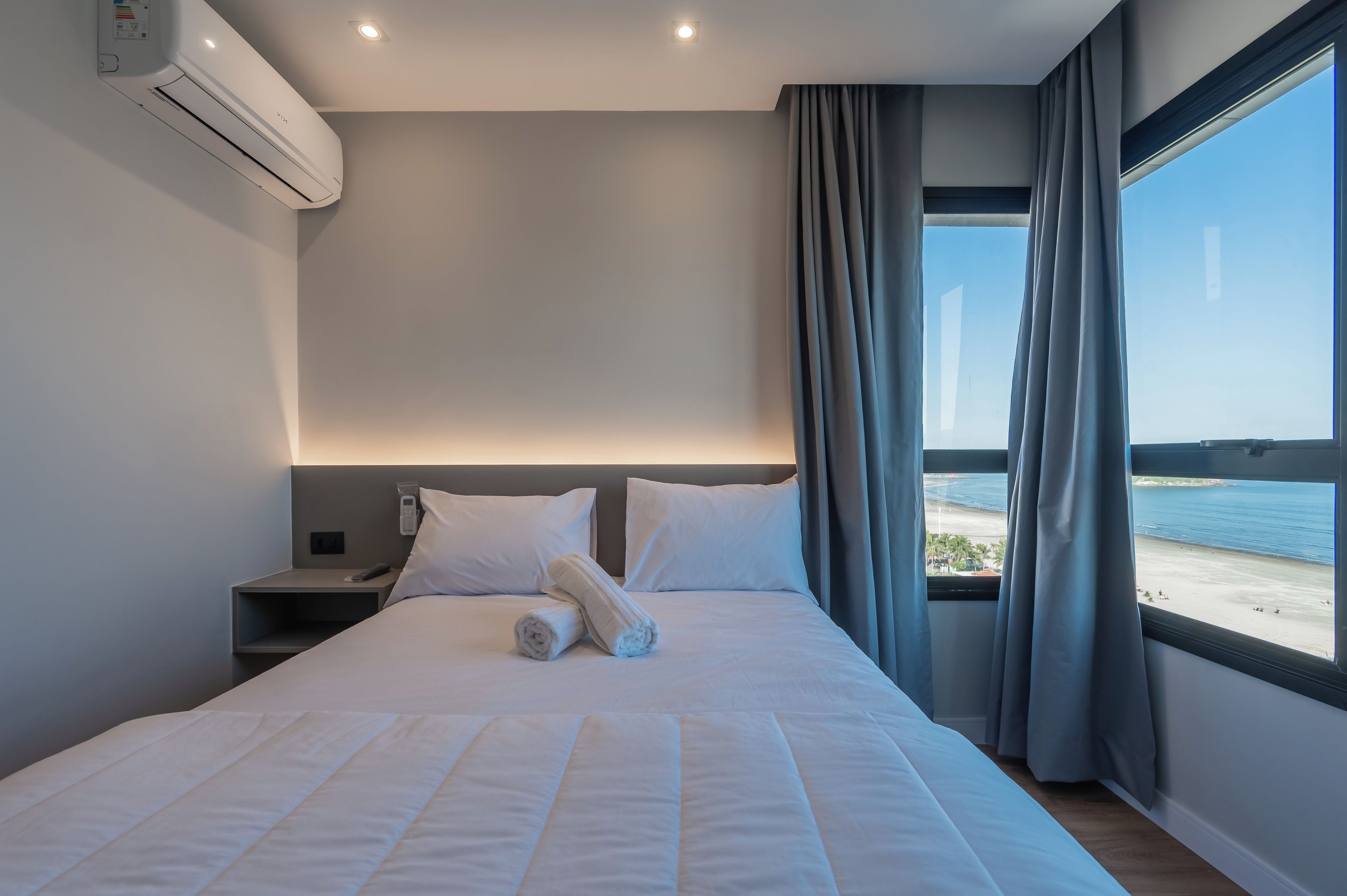 Habitación de hotel con cama, mesita de noche, control remoto, toalla enrollada y vista a la playa a través de una ventana.
