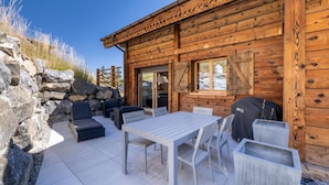 Una cabaña de madera con un área de patio que cuenta con una mesa, sillas, una parrilla y un muro de piedra.