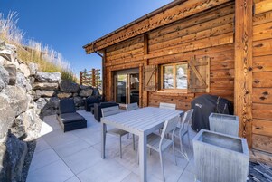 Una cabaña de madera con un área de patio que cuenta con una mesa, sillas, una parrilla y un muro de piedra.