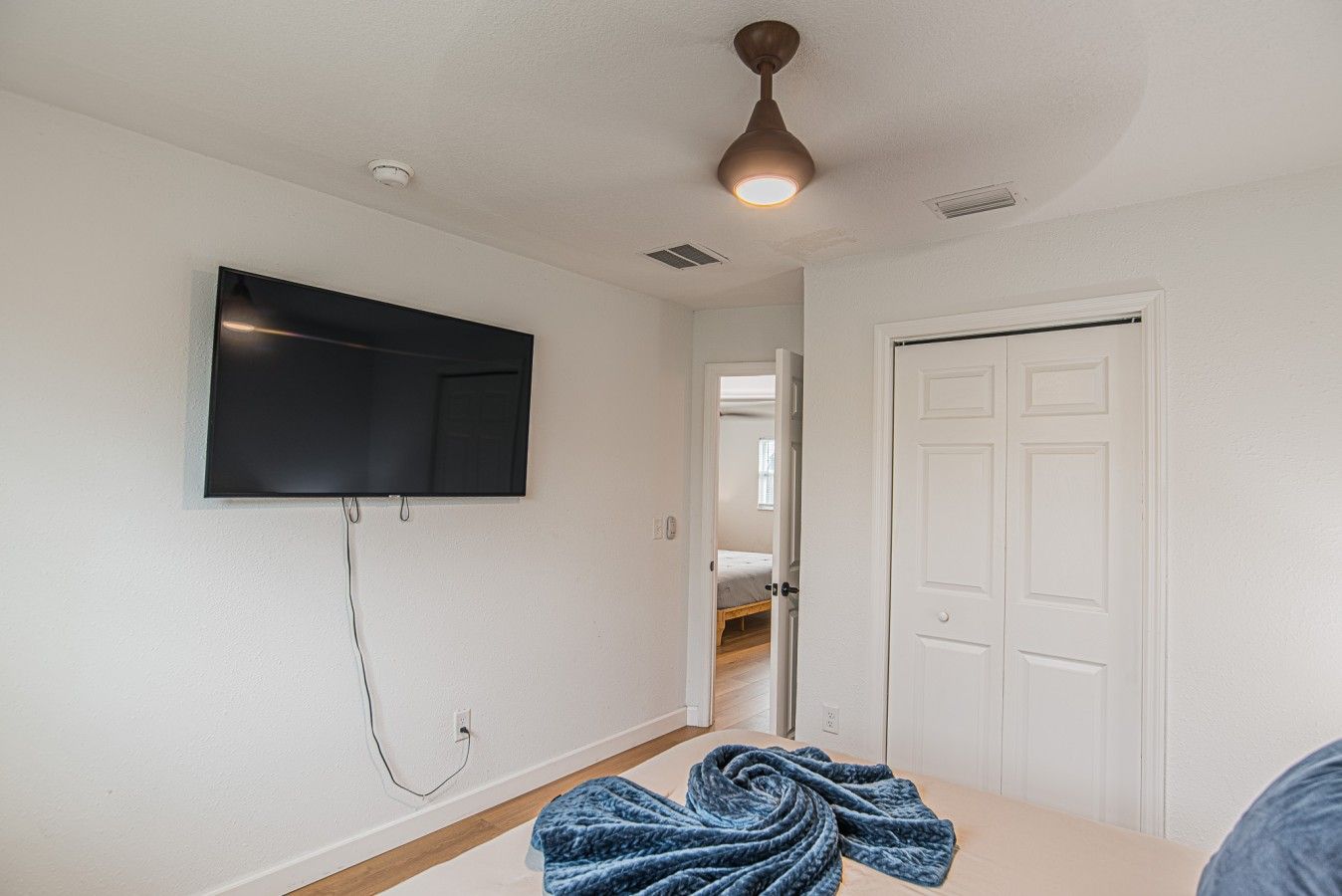 A room with a flat-screen TV mounted on the wall, a ceiling fan, a door, and a bed with a blue blanket.