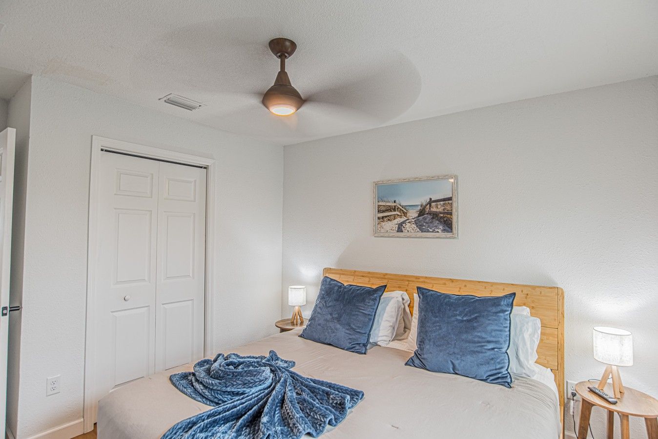 A bedroom with a bed, a ceiling fan, a door, a bedside table with a lamp, and a framed picture on the wall.