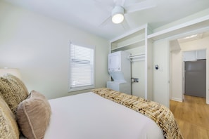 Ein Schlafzimmer mit einem Bett, Deckenventilator, Fenster mit Jalousien sowie eine Waschmaschine und ein Kühlschrank im angrenzenden Raum.