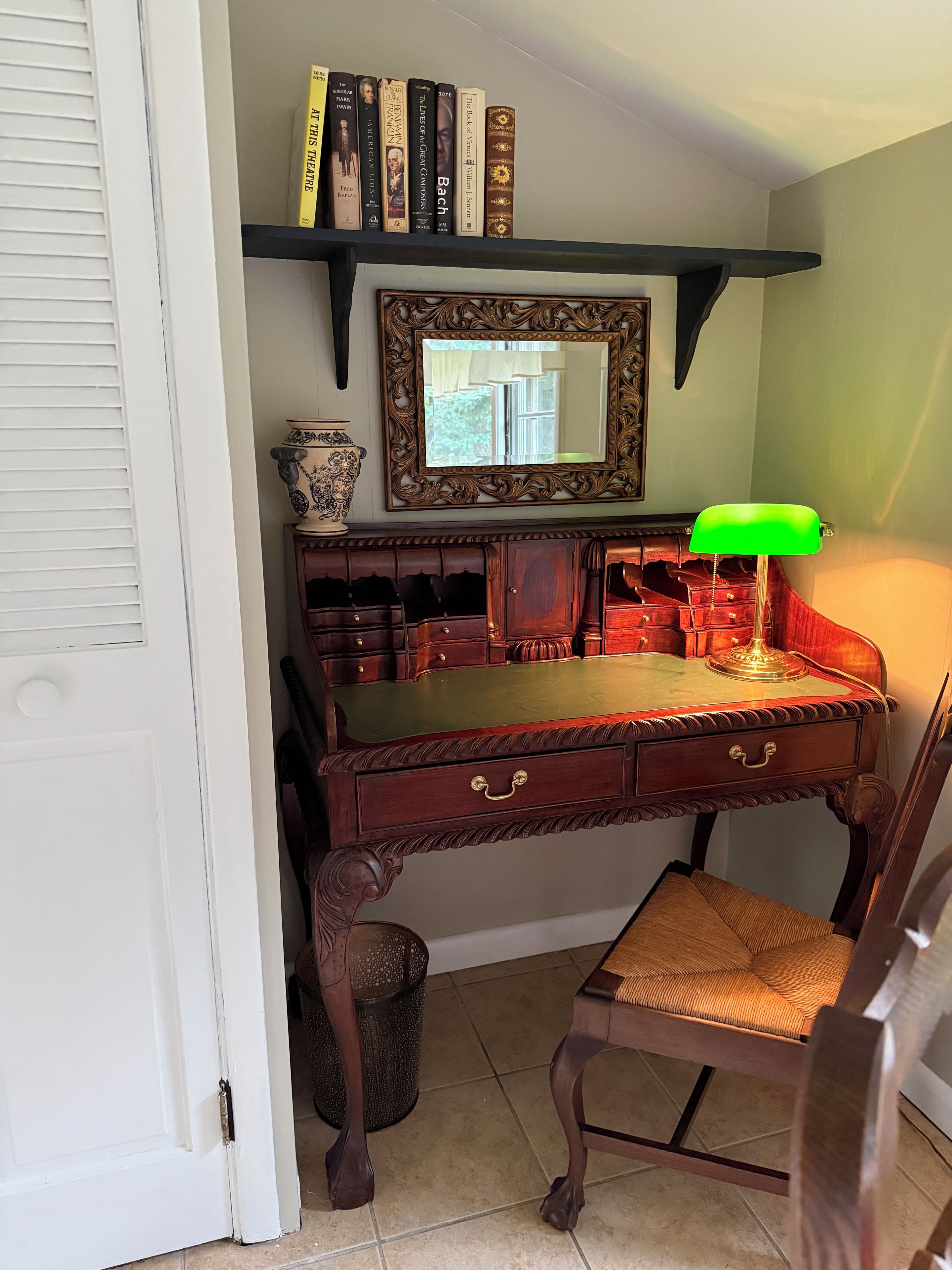 A study room with a wooden desk, a green desk lamp, a chair, a bookshelf with books, a decorative mirror, and a window.