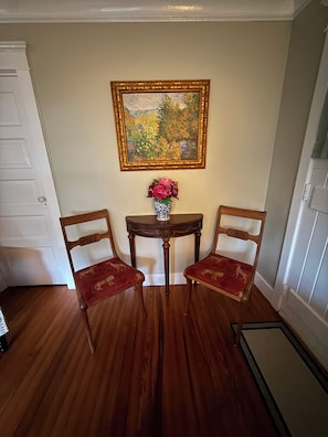 A room with wooden flooring, a white door, a small table with a vase of flowers, and two chairs with red upholstery.