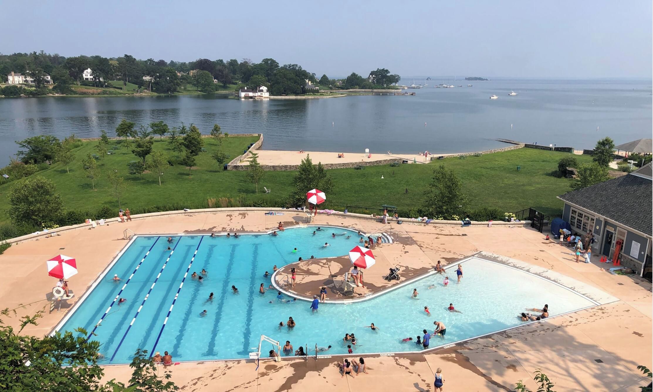 A swimming pool with multiple lanes, surrounded by a sandy area with umbrellas, and a view of a lake with boats.
