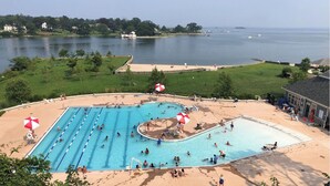 Une piscine avec plusieurs couloirs, entourée d’une zone sablonne agrémentée de parasols, et offrant une vue sur un lac où naviguent des bateaux.