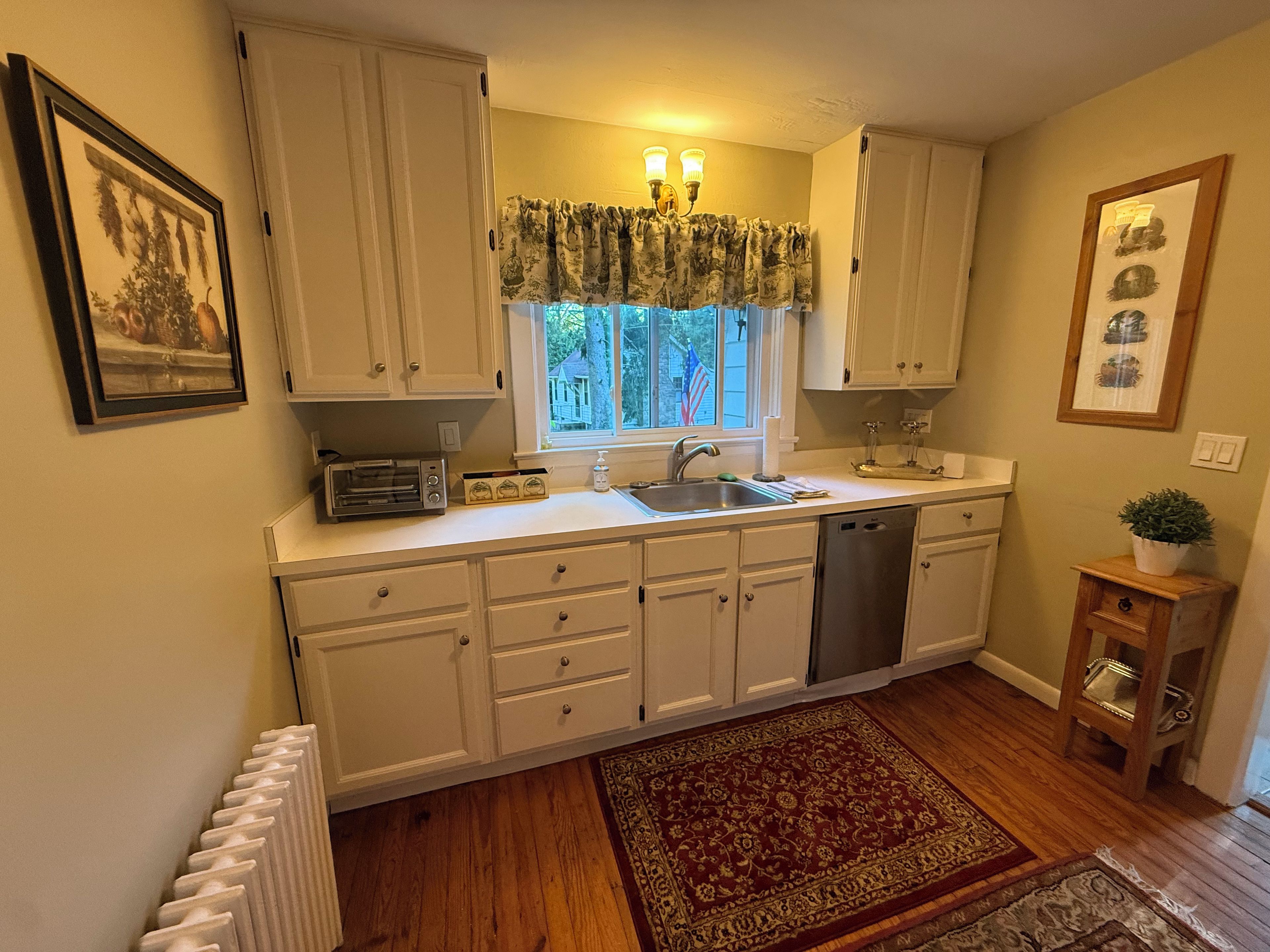 A kitchen with white cabinets, a built-in dishwasher, a window with curtains, and a radiator.
