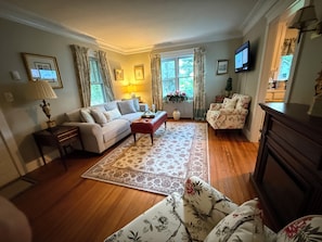 A living room with a sofa, armchair, and a coffee table. There is a fireplace, a television, and a window with curtains.