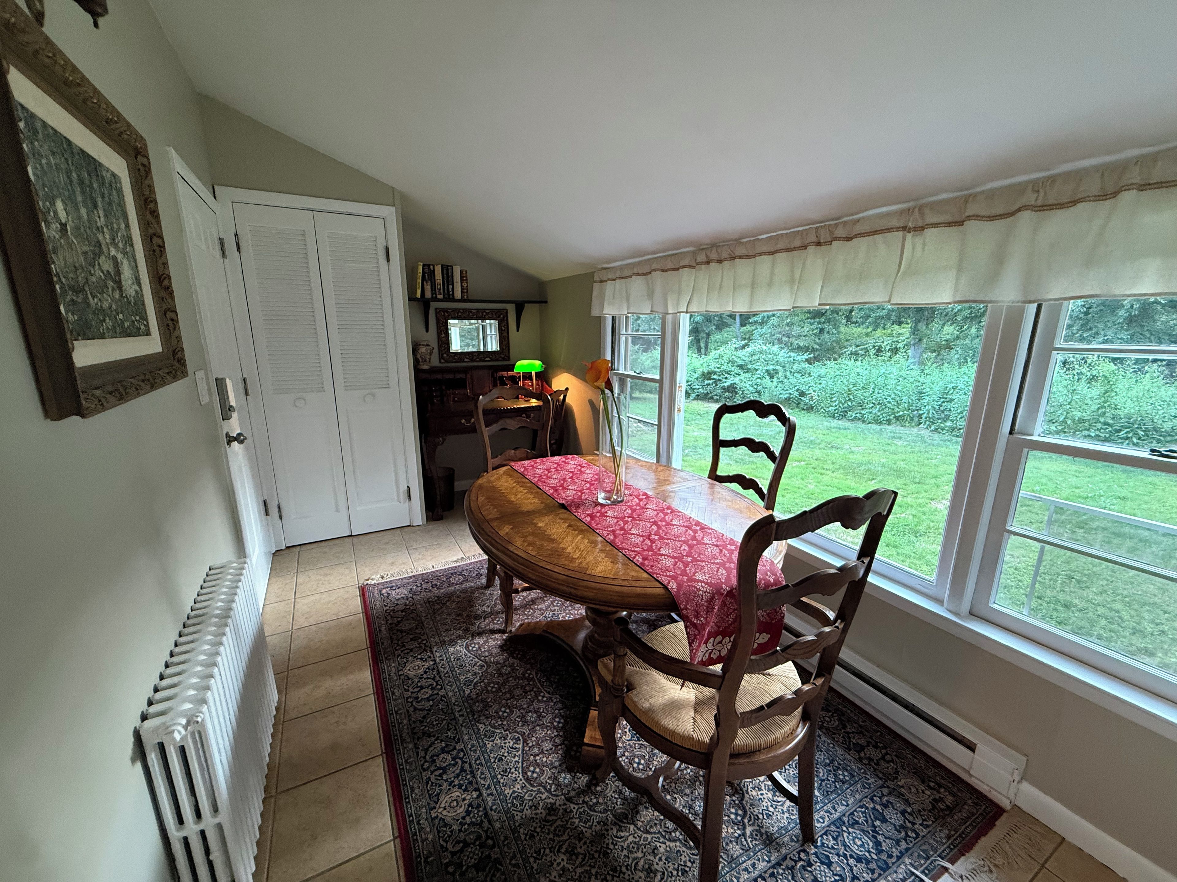 A dining area with a table, chairs, a radiator, a window with a view of greenery, and a door.