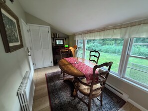 Un comedor con una mesa, sillas, un radiador, una ventana con vistas a follaje y una puerta.