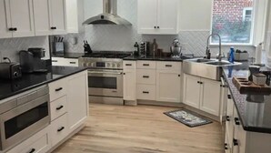 Una cocina con armarios blancos, campana extractora de acero inoxidable, horno empotrado, un lavamanos y una ventana con vista a un edificio de ladrillo.