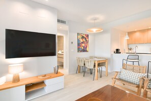 A modern living room with a flat-screen TV, a dining area with a table and chairs, and a kitchen with wooden cabinets and a pendant light. - Los Colibríes by H - Isla Canela Golf GLHC7023P98 (Ayamonte)