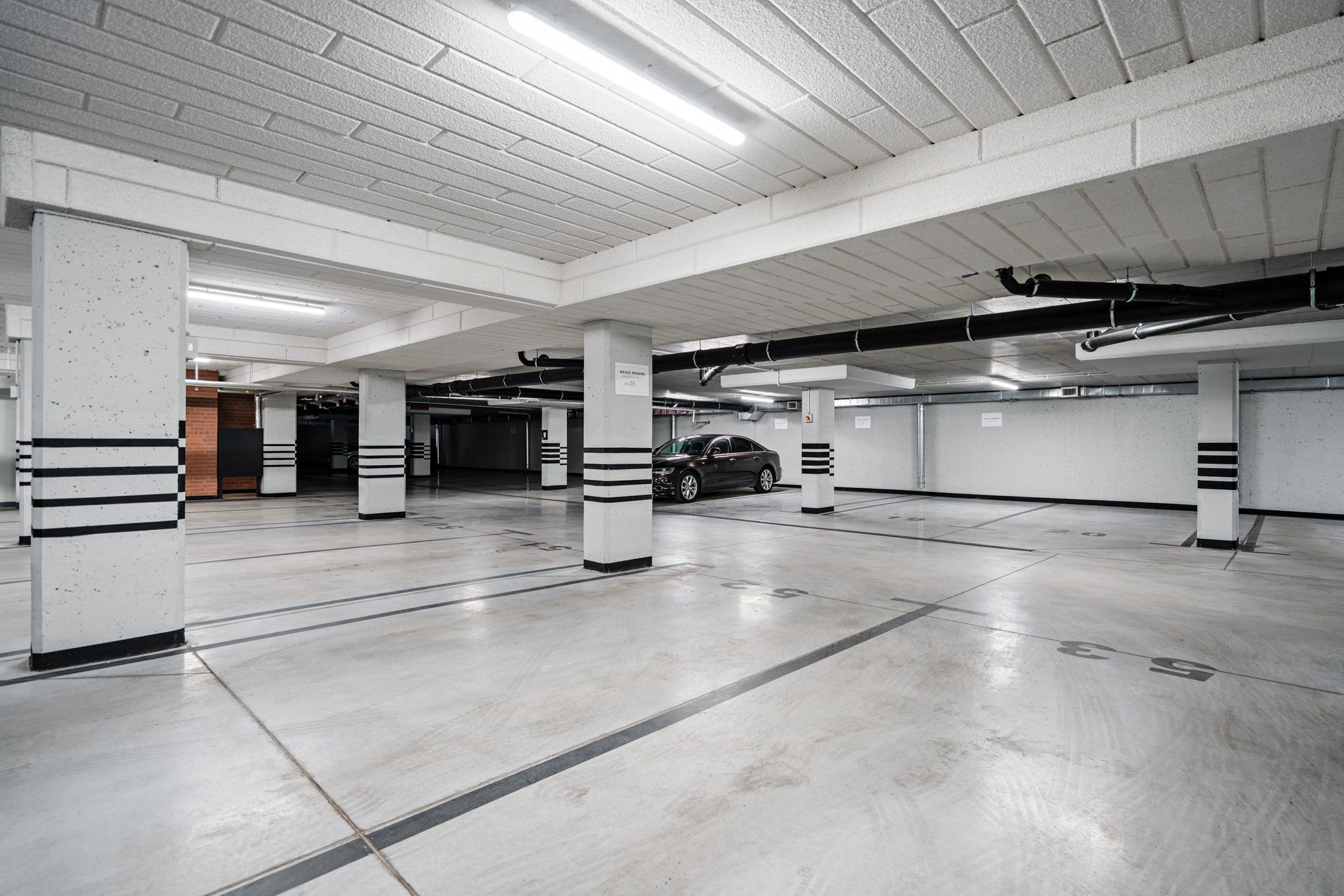 Un aparcamiento de varios niveles con pilares de hormigón, un coche estacionado en una de las plazas y luces en el techo.