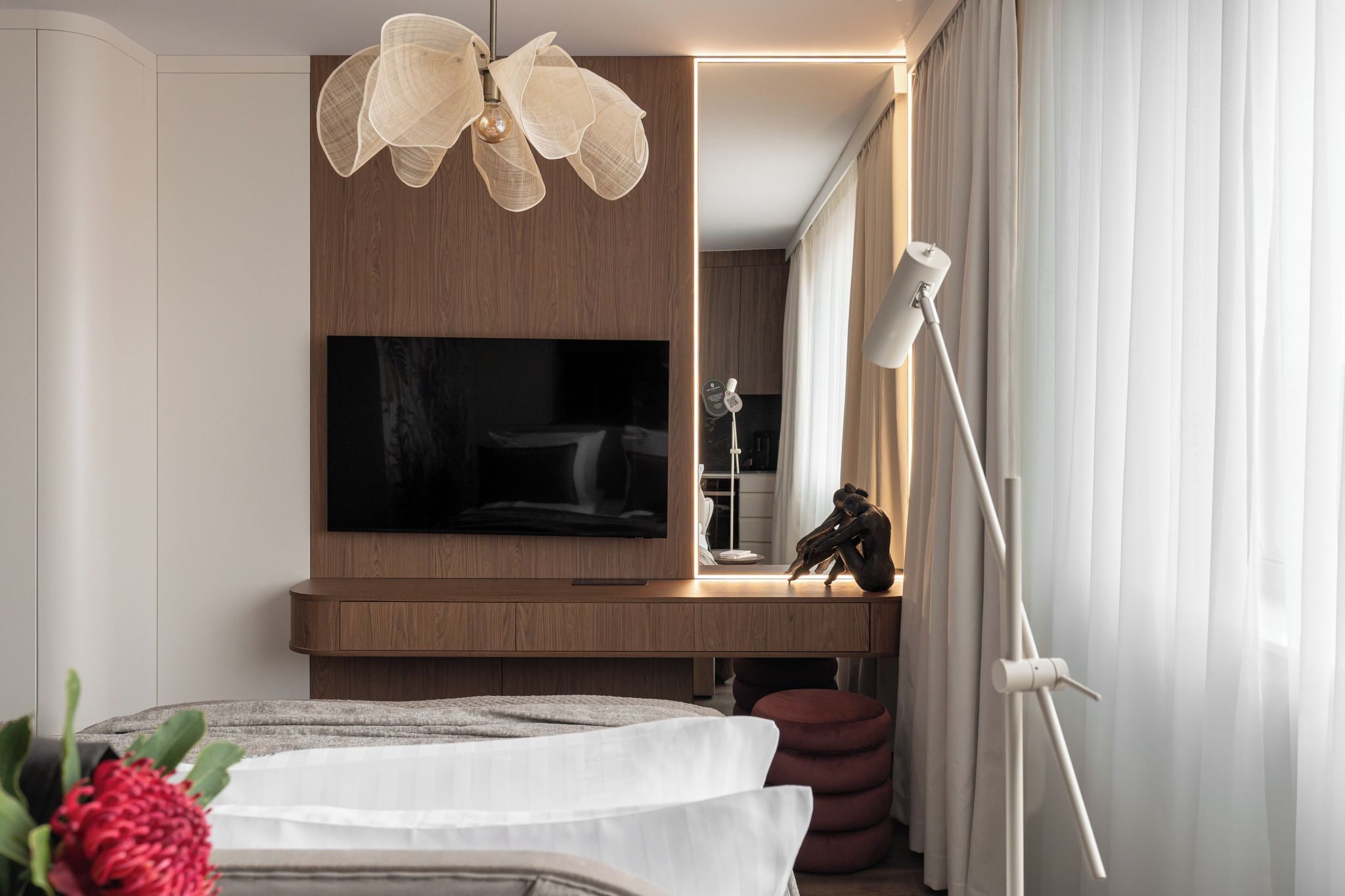 Ein modernes Schlafzimmer mit einem großen Flachbildfernseher, einer Holzkommode, einer Stehlampe und einem Bett mit weißer Bettdecke.