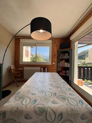 A room with a large window, a table with a leaf-patterned tablecloth, a bookshelf, and a chair. - Le Narcisse—Mountain View, Sleeps 6, Quiet, Ski Shuttle (Villard-de-Lans)