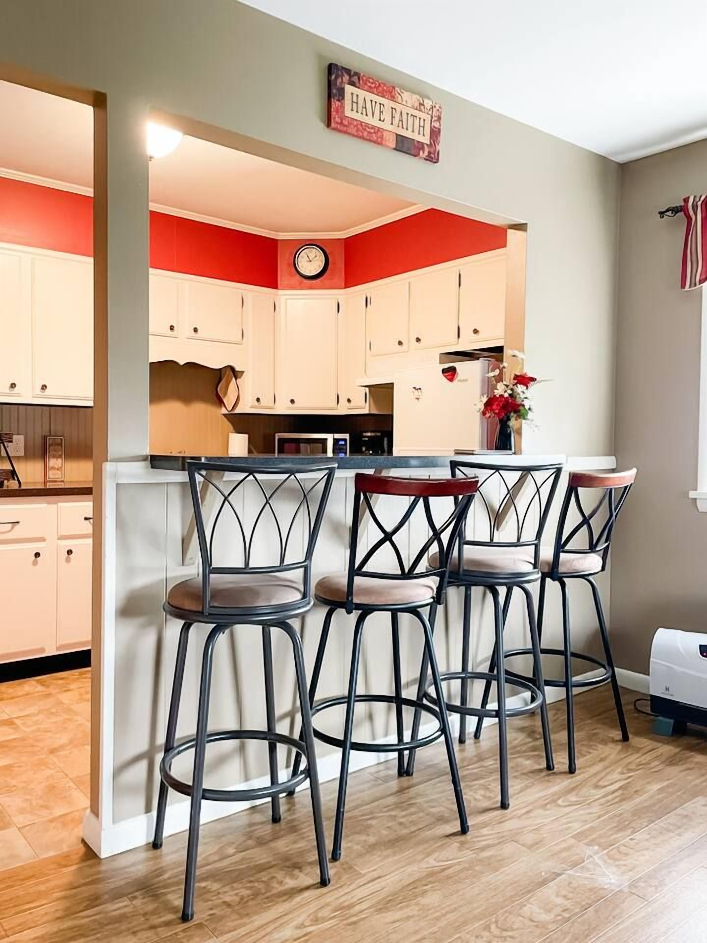 Une cuisine avec un mur d’accent rouge, une horloge et un panneau indiquant « Ayez foi ». On y trouve trois tabourets de bar et un four à micro-ondes.