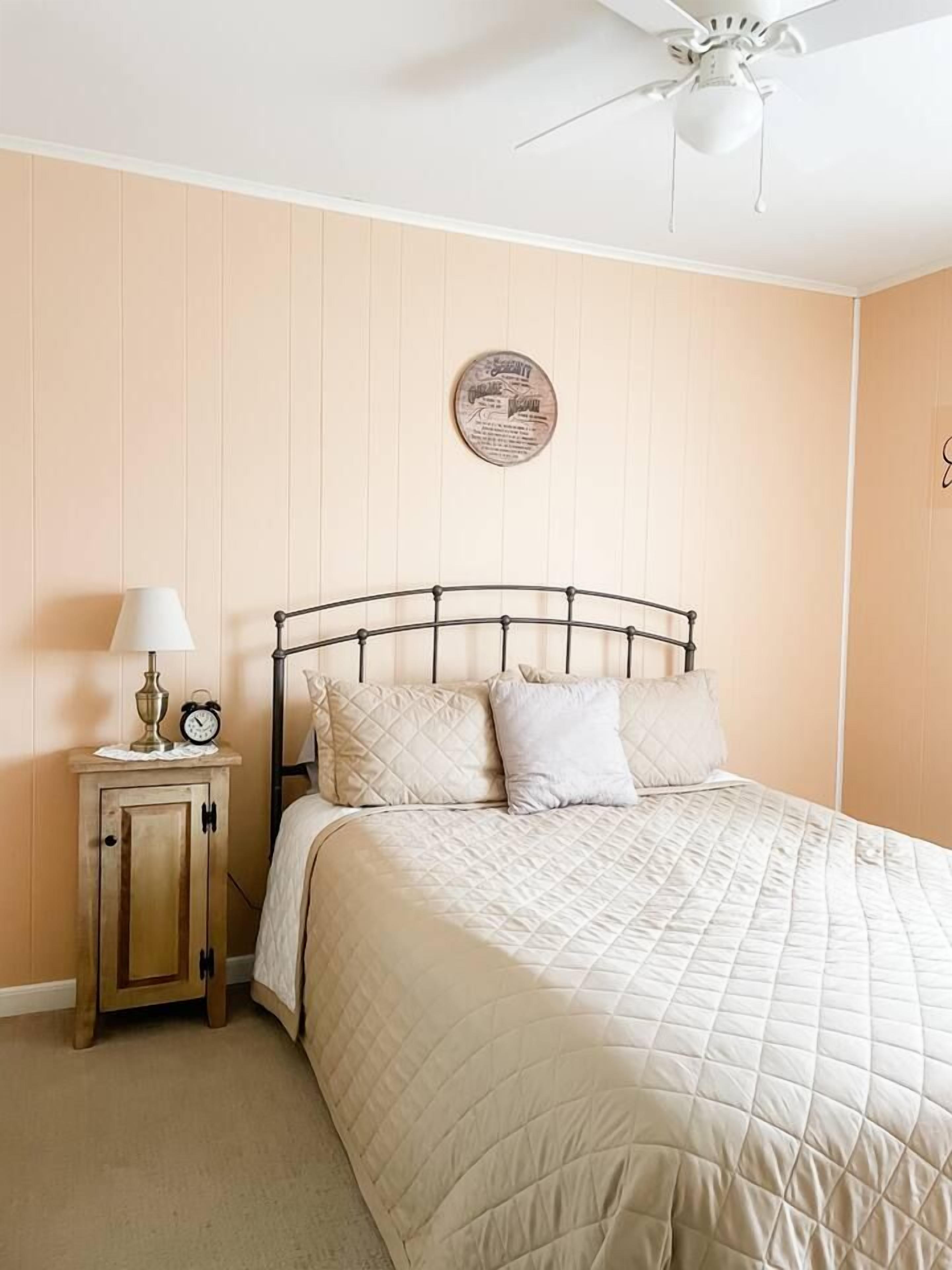 A bedroom with a bed, a nightstand, a lamp, a clock, and a ceiling fan.