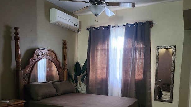 Un dormitorio con cama, ventilador de techo, aire acondicionado, mesita de noche, espejo y baldosas de suelo estampadas.