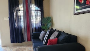 Una sala de estar con un sofá negro, pisos de baldosas con diseño, una ventana con cortinas y un cuadro en la pared.