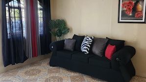 Una habitación con ventilador de techo, un sofá negro con varios cojines, una planta en maceta y un cuadro colgado en la pared.