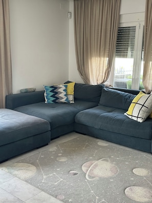Ein winkelförmiges Ecksofa mit gemusterten Kissen, ein grauer Teppich mit Kreis-Motiven und ein Fenster mit Jalousien.