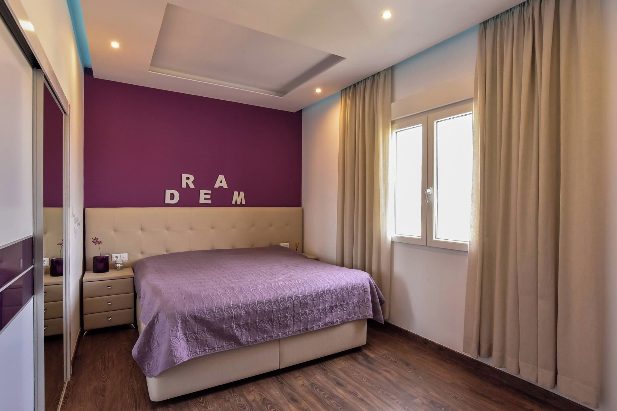 Un dormitorio con cama, mesita de noche, cómoda, ventana con cortinas y pared con letras.