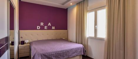Un dormitorio con cama, mesita de noche, cómoda, ventana con cortinas y pared con letras.