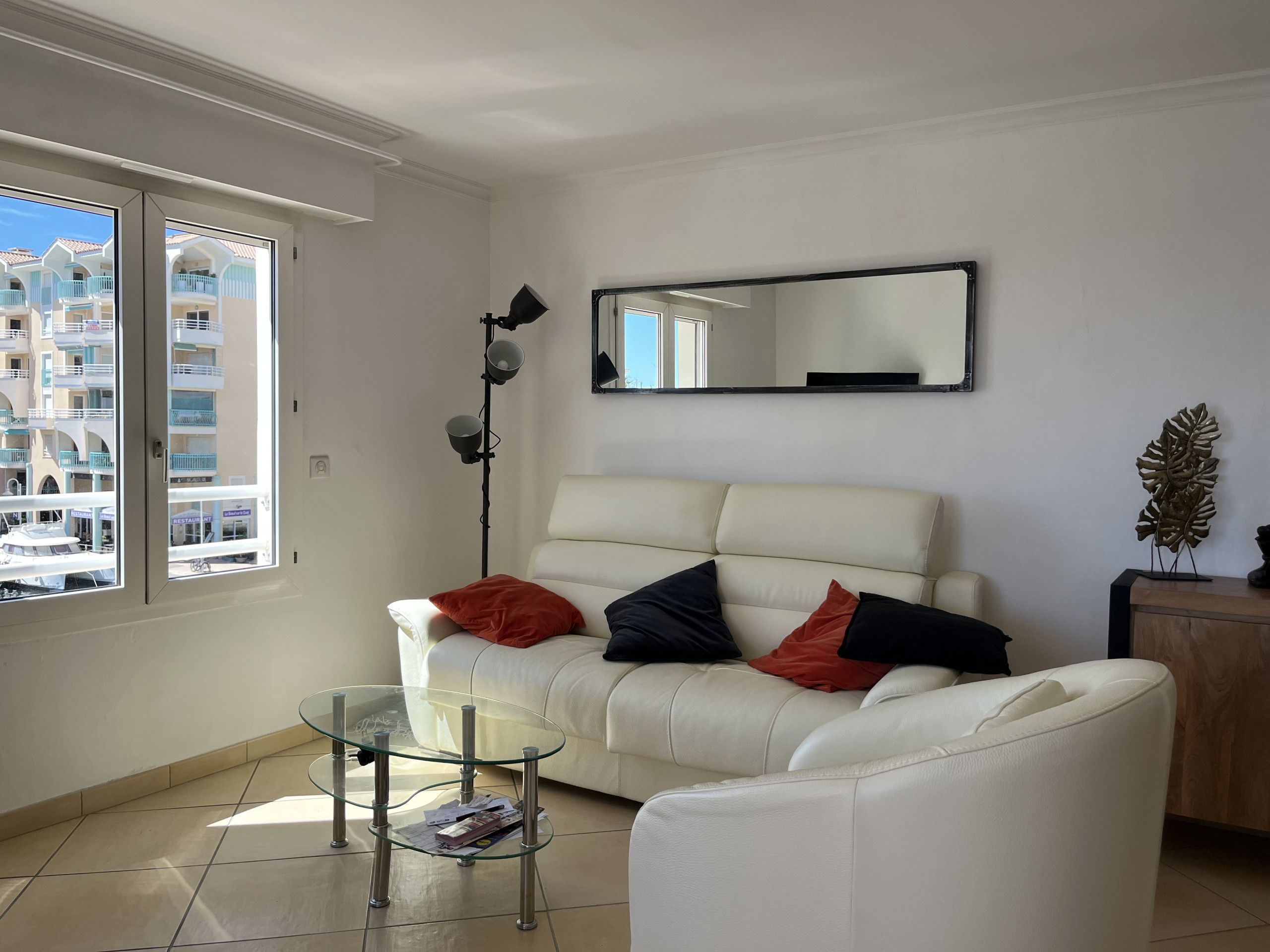 A living room with a white sofa, a glass coffee table, a floor lamp, and a view of buildings through a window.