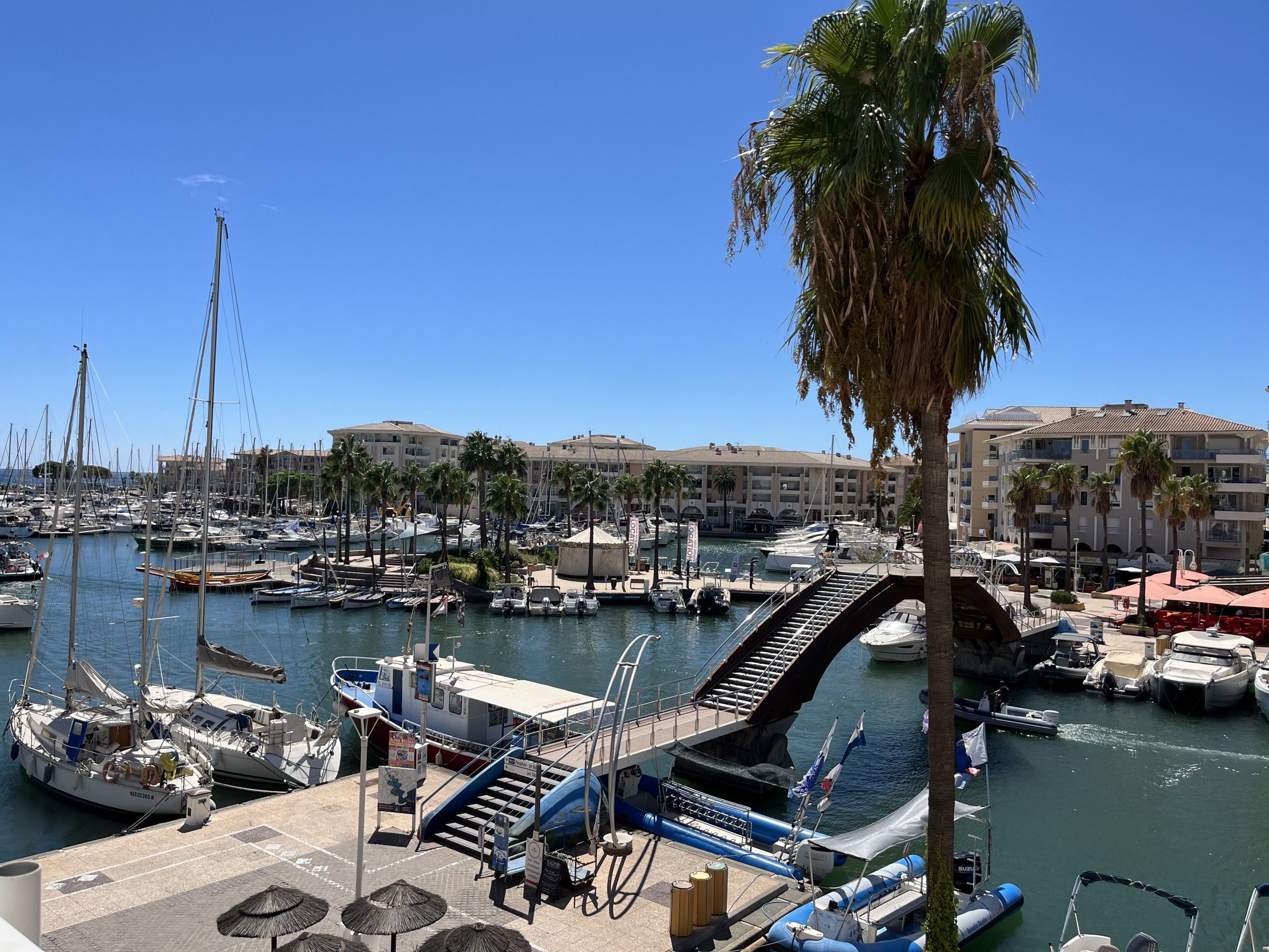 A marina with boats docked, a pedestrian bridge, and buildings in the background.