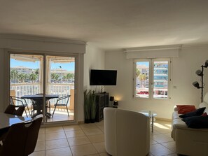 A living room with a TV, a sofa, a dining table with chairs, and a balcony with a view of buildings and palm trees. - Port Fréjus: magnificent 1-bedroom apartment with a harbor view, sleeping 4; lively area, air-conditioned, with Wi-Fi and parking. (Fréjus)