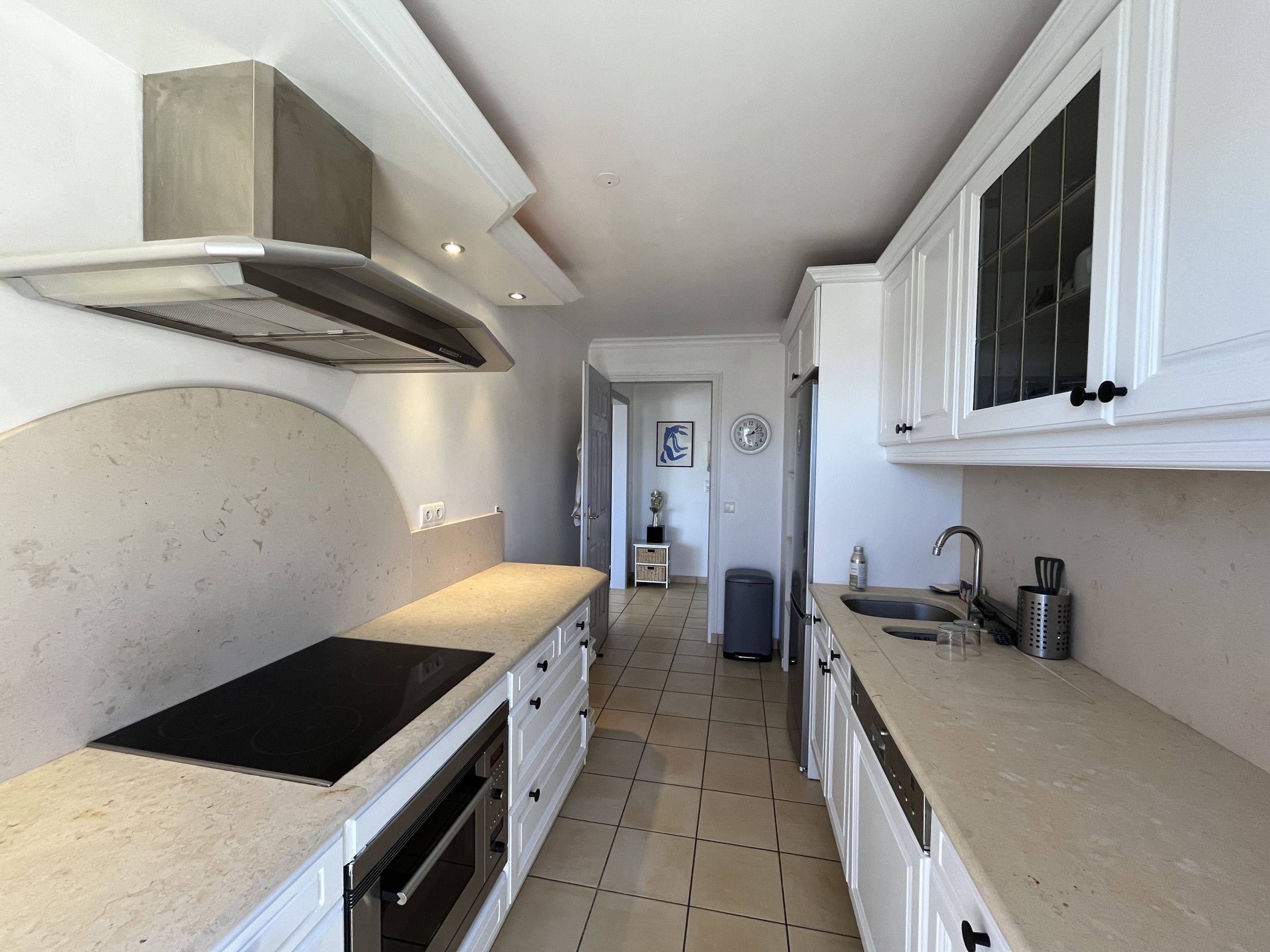A narrow kitchen with white cabinets, a stainless steel range hood, a built-in oven, and a tiled floor.