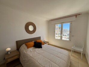 A bedroom with a bed, a bedside table, a lamp, a chair, a window with a view of buildings, and a wall-mounted mirror. - Port Fréjus: magnificent 1-bedroom apartment with a harbor view, sleeping 4; lively area, air-conditioned, with Wi-Fi and parking. (Fréjus)