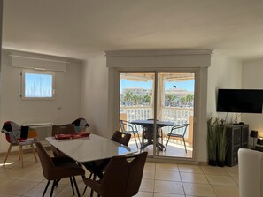 Un coin repas avec une table et des chaises, un téléviseur fixé au mur et un balcon offrant une vue sur les bâtiments et les palmiers.
