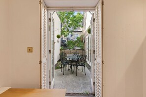 Ein schmaler Außenbereich mit einem Tisch und Stühlen, der zu einem Garten mit einer Bank und hängenden Pflanzen führt.
