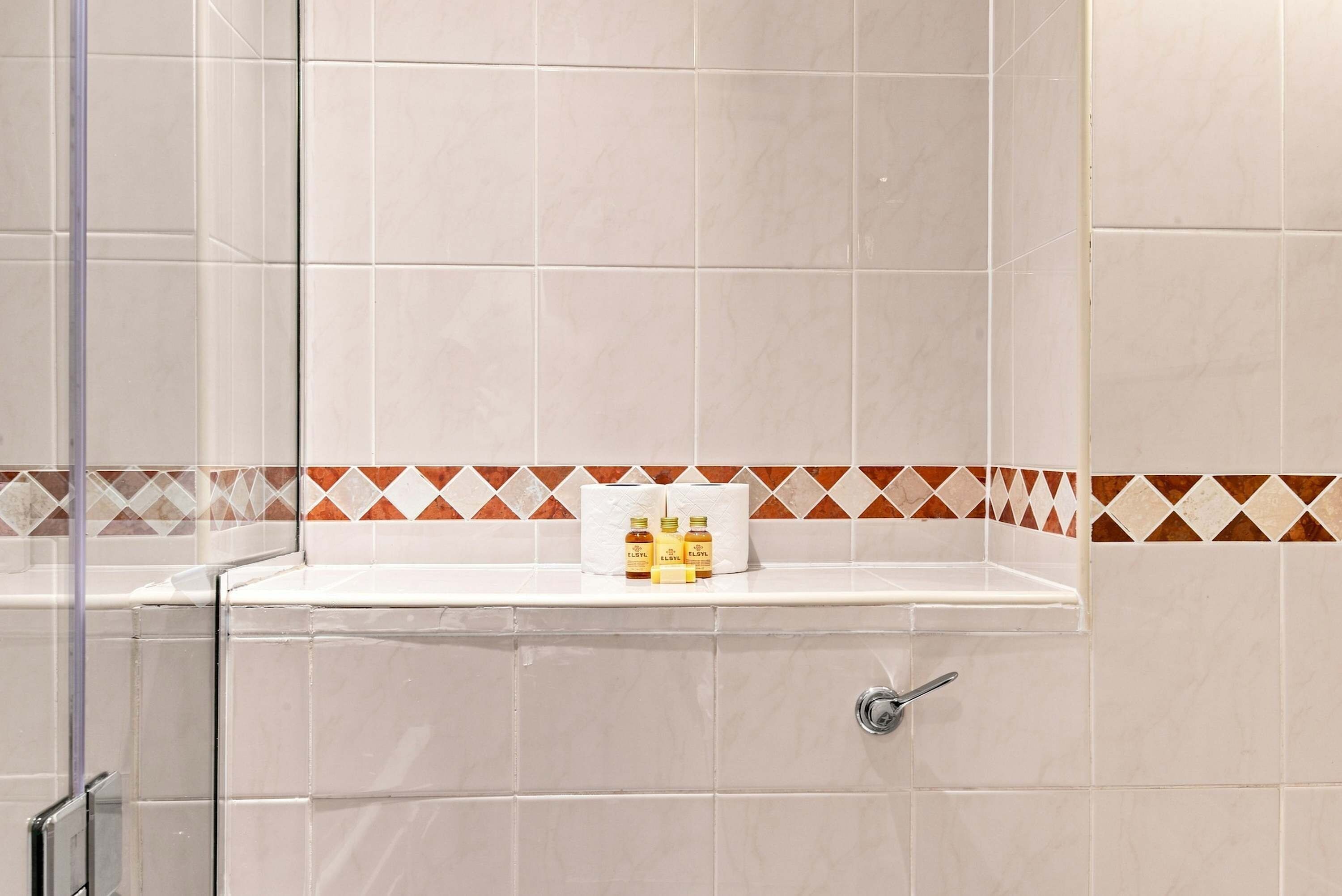 Une salle de bain carrelée de blanc, avec une douche et une étagère où sont rangés les produits de toilette.