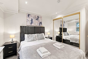 Quarto moderno com uma cama grande, mesas de cabeceira com abajures, um espelho grande e arte abstrata na parede.