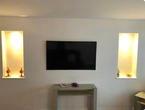 A room with a flat-screen TV mounted on the wall, flanked by two wall-mounted light fixtures with shelves. - Apartment Sleeps 3 With Wifi (Alexandria)