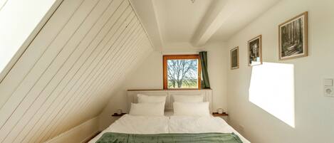 A bedroom with a sloped ceiling, a bed with white bedding, a window with a view of trees, and two framed pictures on the wall.