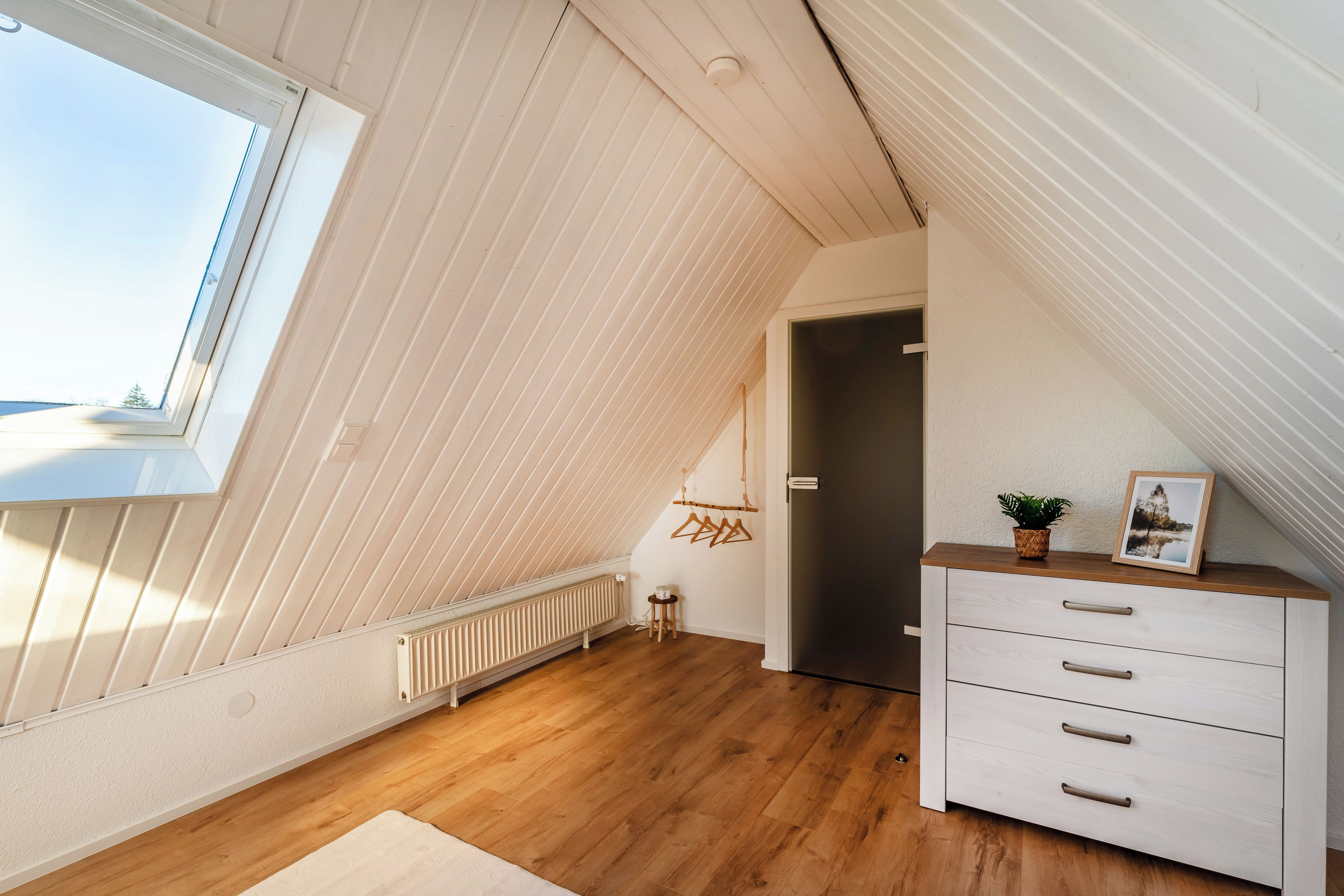 Habitación con techo inclinado, un radiador, suelo de madera, un aparador blanco con cajones, una pequeña mesa con una planta y una ventana con vistas.