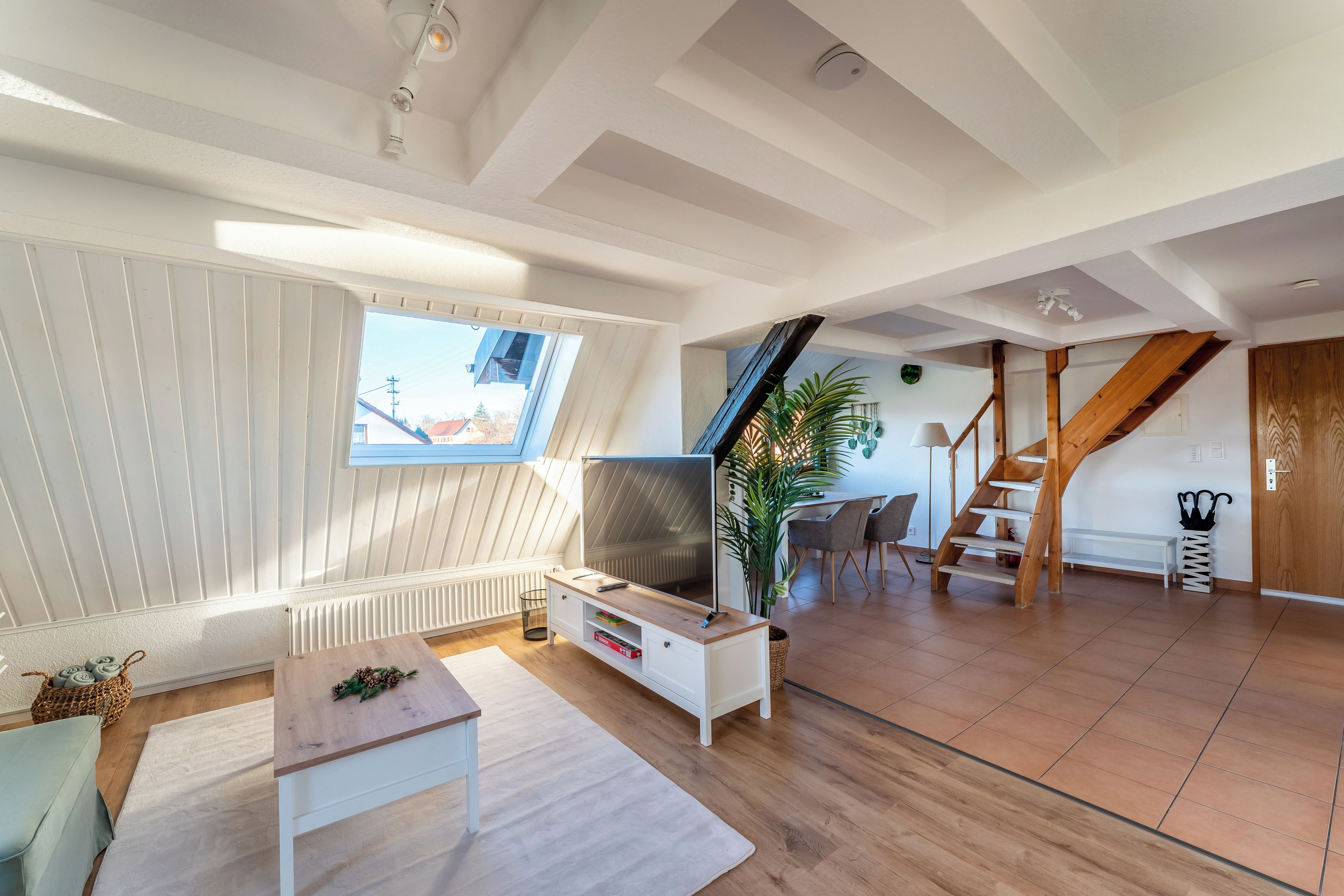 A spacious loft with a high ceiling, a large window, a TV, a wooden staircase, and a dining area.