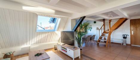 A spacious loft with a high ceiling, a large window, a TV, a wooden staircase, and a dining area.