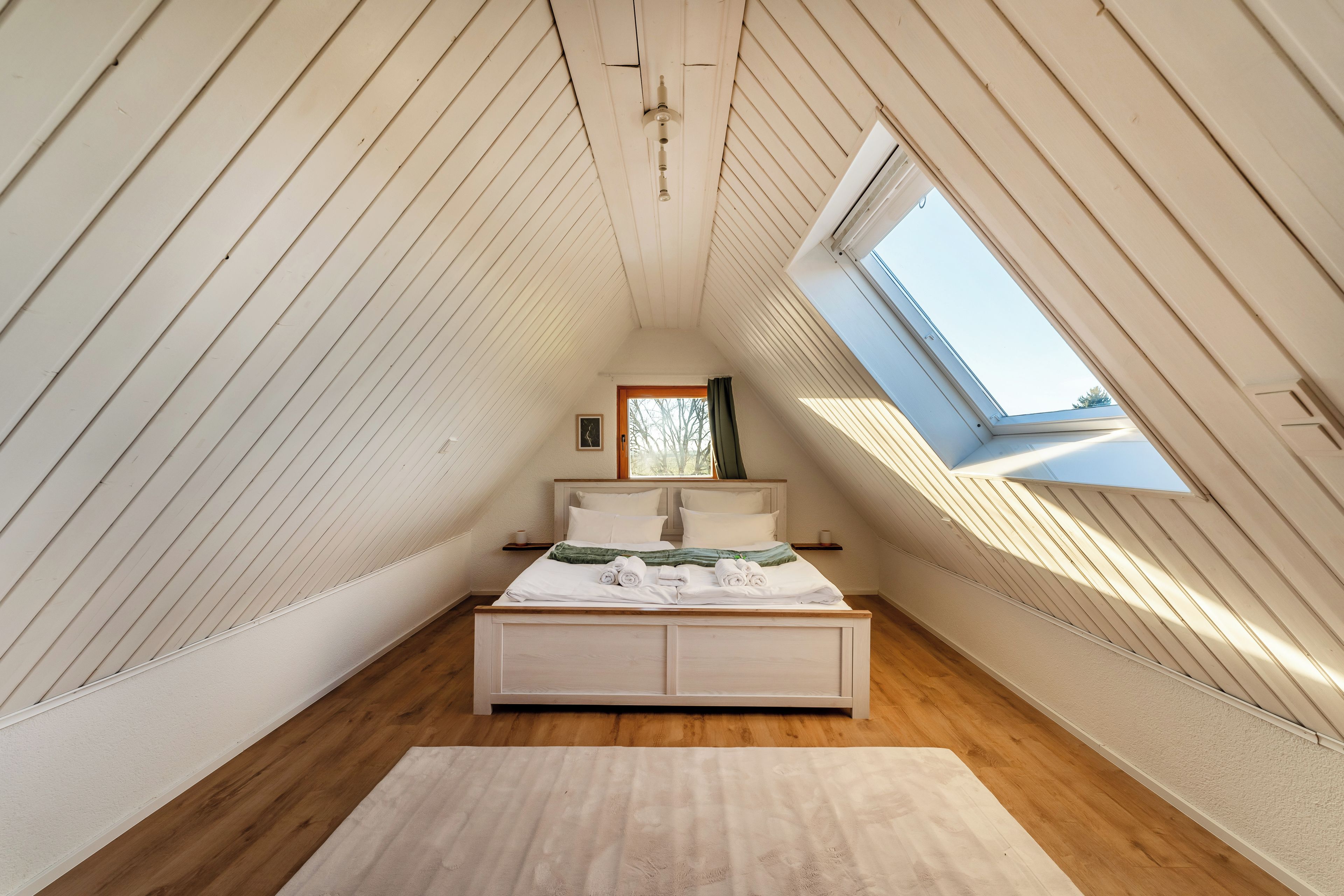 A bedroom with a sloped ceiling, a bed with white bedding, a small bedside table, and a window.