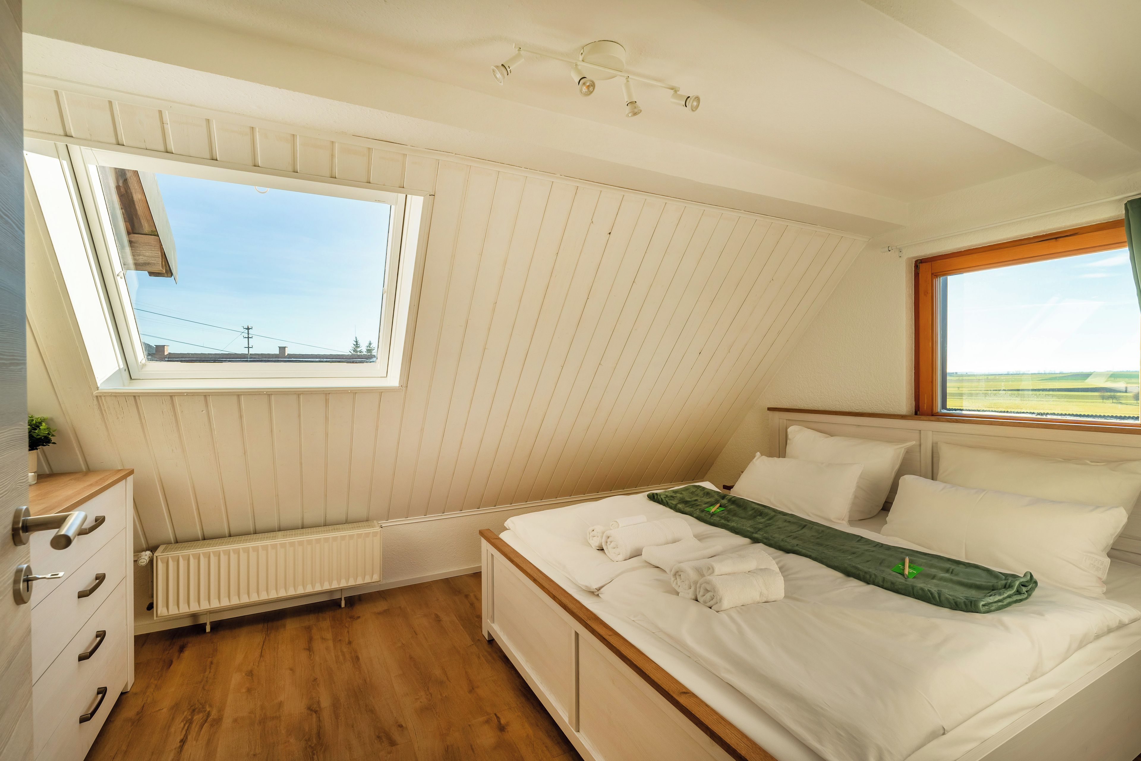 Une chambre avec un plafond en pente, une fenêtre donnant sur un beau paysage, un lit avec du linge de maison blanc et un radiateur.