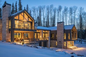 Un chalet moderne à plusieurs étages, doté d’un grand balcon, d’une façade en pierre et surplombant une forêt enneigée.