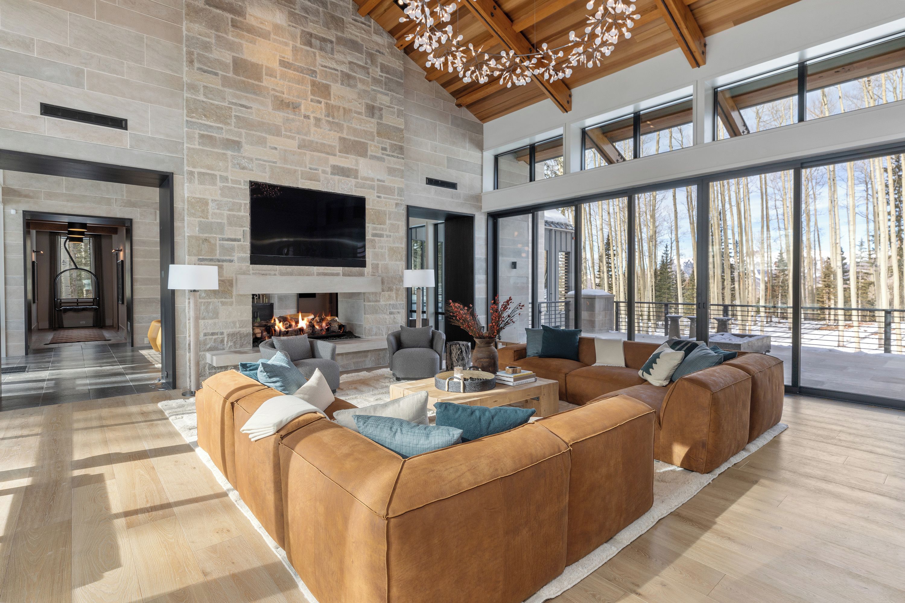 A spacious living room with a stone fireplace, leather sofas, and large windows offering a view of trees.