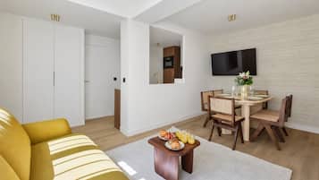 Un salon moderne avec un canapé, une table à manger et des chaises, un téléviseur à écran plat fixé au mur, et un coin cuisine à l’arrière-plan.