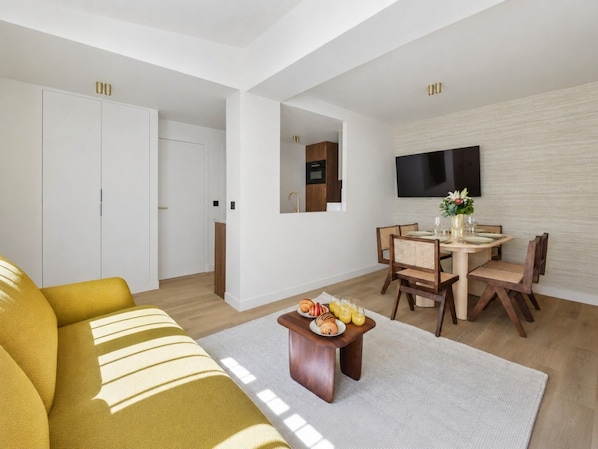 A modern living room with a sofa, a dining table with chairs, a flat-screen TV mounted on the wall, and a kitchen area in the background. - Lavie Maison – Hot Tub & AC – Central Paris (Paris)